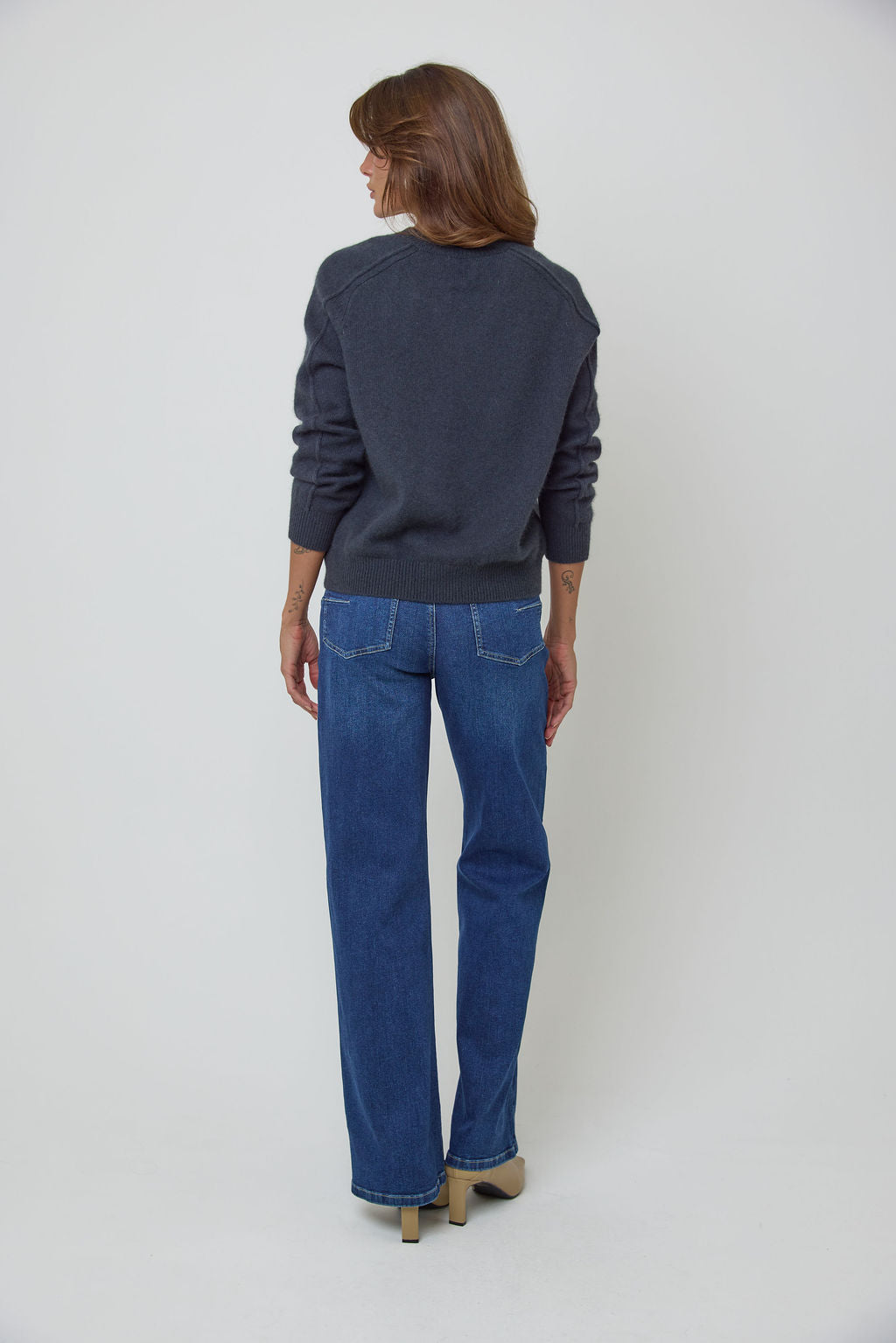 A woman modelling a charcoal blue V-neck knit wool sweater and blue straight-leg jeans against a light grey studio background.