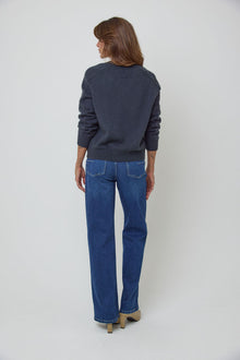 A woman modelling a charcoal blue V-neck knit wool sweater and blue straight-leg jeans against a light grey studio background.