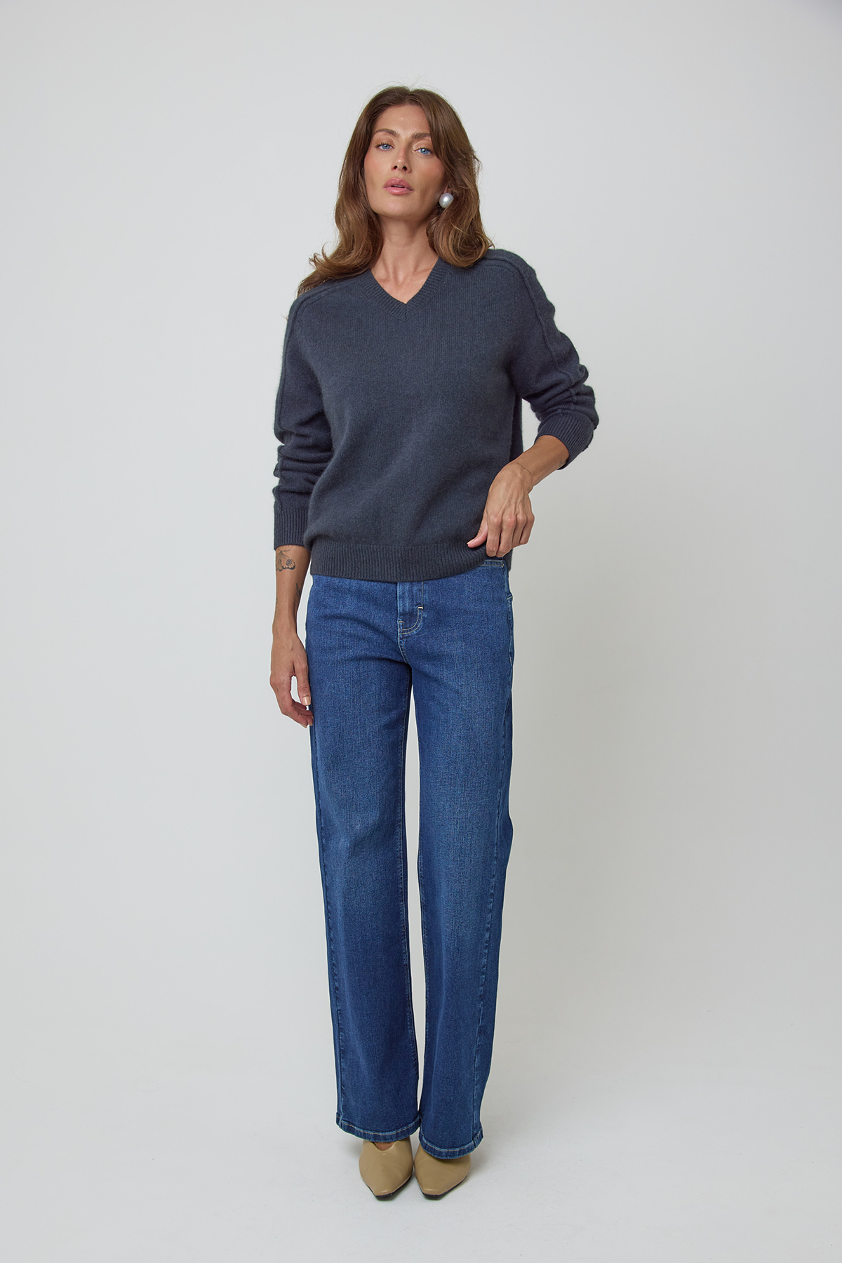 A woman modeling a charcoal blue V-neck knit wool sweater and blue straight-leg jeans against a light gray studio background.