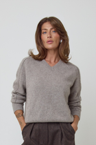 A woman wearing a light grey/brown v-neck cashmere knit sweater and dark brown wool trousers, standing against a plain light grey studio background.