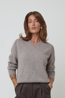 A woman wearing a light grey/brown v-neck cashmere knit sweater and dark brown wool trousers, standing against a plain light grey studio background.