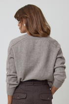 A woman wearing a light grey/brown v-neck cashmere knit sweater and dark brown trousers, standing against a plain light grey studio background.