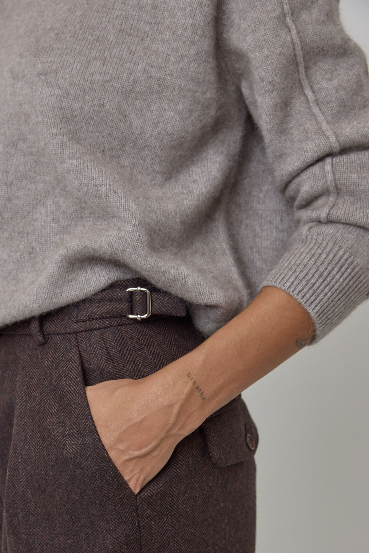 Close-up of a person wearing a light grey/brown cashmere knit sweater and dark brown textured wool trousers with adjustable waist details.