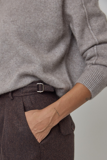 Close-up of a person wearing a light grey/brown cashmere knit sweater and dark brown textured wool trousers with adjustable waist details.