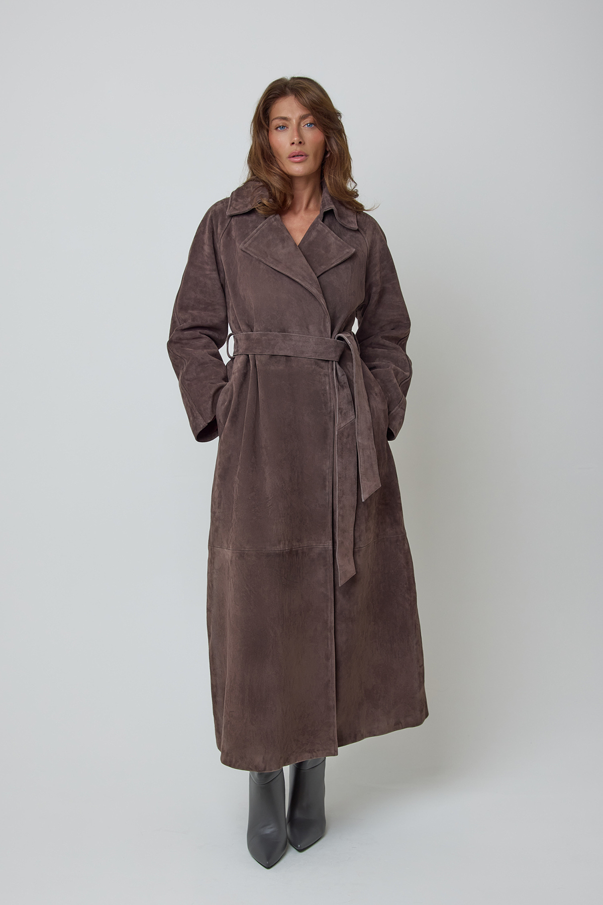 A woman modelling a long, dark brown real genuine suede belted long trench coat against a plain light grey studio background.