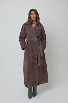 A woman modelling a long, dark brown real genuine suede belted long trench coat against a plain light grey studio background.