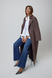 A woman sits on a chair wearing a long brown suede trench coat over a white button-up cotton shirt, blue wide leg jeans, and dark pointed heels against a light grey studio background.