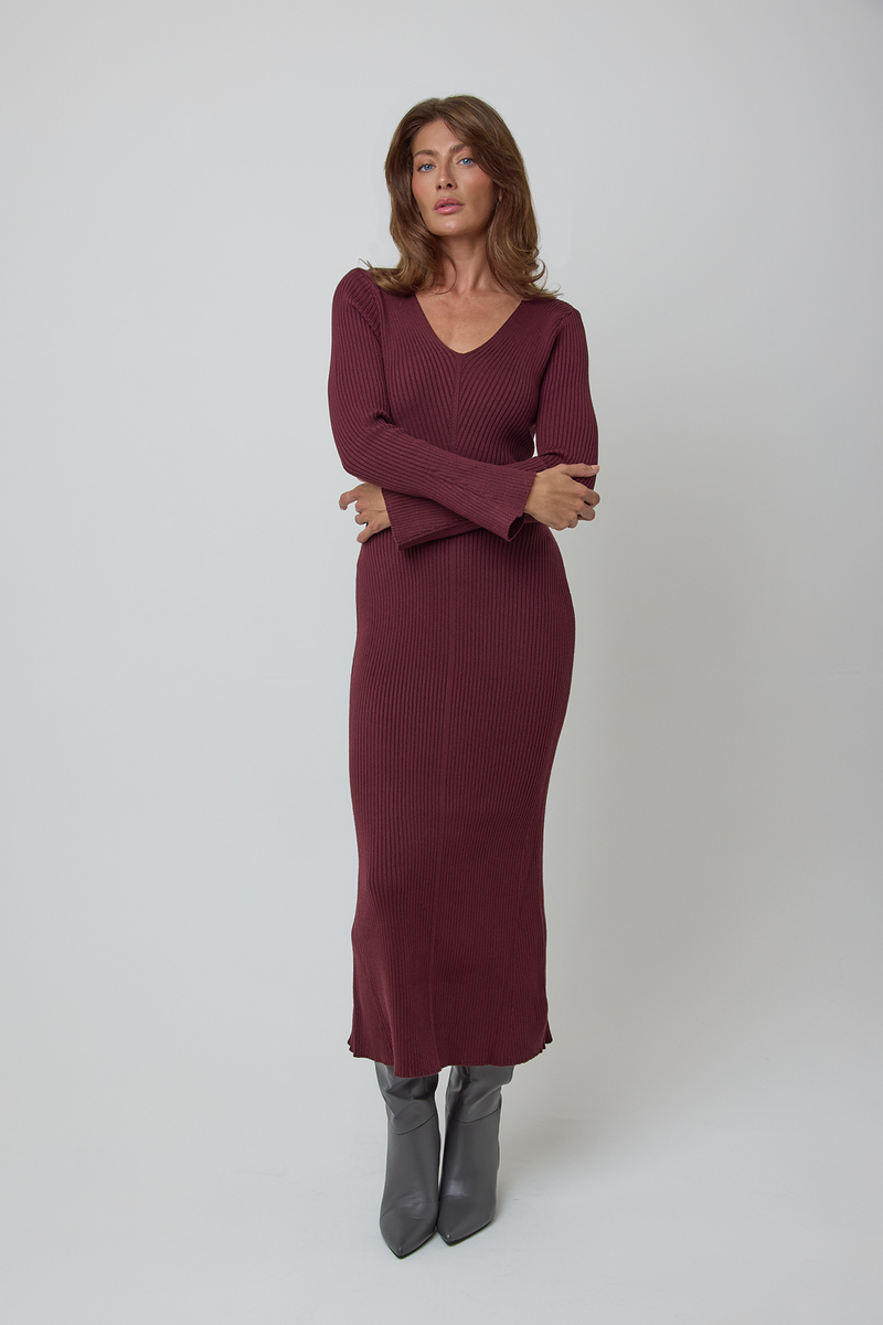 A woman modelling a ribbed long-sleeve wool midi dress in burgundy, paired with dark grey ankle boots, against a light grey studio background.