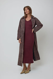 A woman modelling a long brown genuine suede coat over a ribbed burgundy wool midi dress, paired with beige pointed-toe heels, against a light grey studio background.