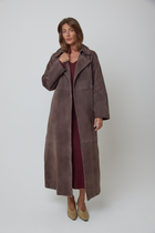 A woman modelling a long, dark brown real suede coat over a burgundy wool dress and beige heels, photographed against a plain light grey studio background.