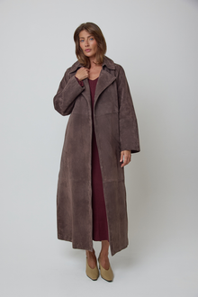 A woman modelling a long, dark brown real suede coat over a burgundy wool dress and beige heels, photographed against a plain light grey studio background.