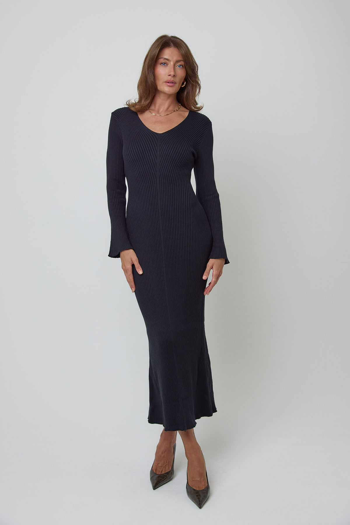 A woman modeling a long-sleeve, ribbed wool knit midi dress in black, paired with dark pointed-toe heels, against a light gray studio background.